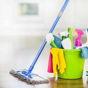Bucket with cleaning items on blurry modern kitchen background. Washing brush and spray set with copy space.